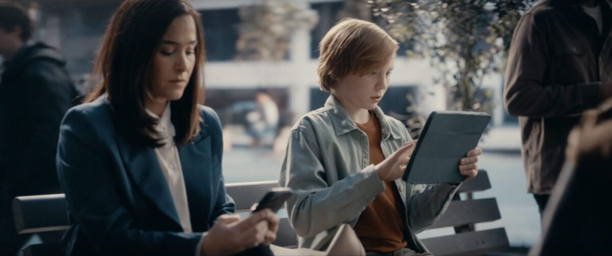 A mom sitting next to her son on a park bench. She is scrolling on her phone while he plays a game on his tablet.